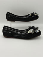 A pair of black woven flat shoes with decorative floral embellishments and rhinestone accents on the toes, displayed on a light gray background with one shoe on a clear acrylic block.