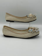 A pair of beige crochet flat shoes with decorative bows and rhinestone embellishments on the toes, displayed on a light gray background with one shoe on a clear acrylic block.
