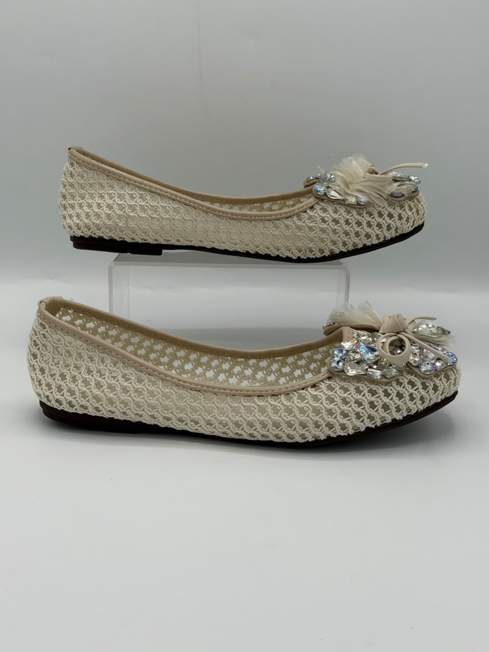 A pair of beige crochet flat shoes with decorative bows and rhinestone embellishments on the toes, displayed on a light gray background with one shoe on a clear acrylic block.