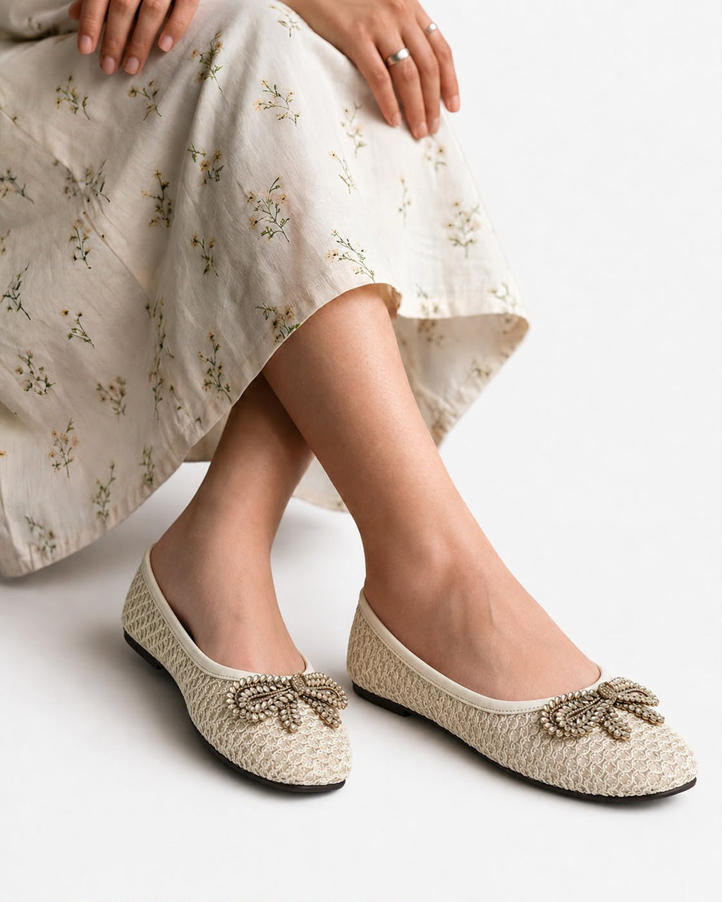 A person wearing woven beige ballet flats with jeweled bow details, paired with a light cream floral skirt, against a clean white background.