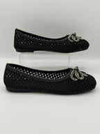 A pair of black knit ballet flats with silver rhinestone bow embellishments, displayed on a clear acrylic stand against a light gray background.