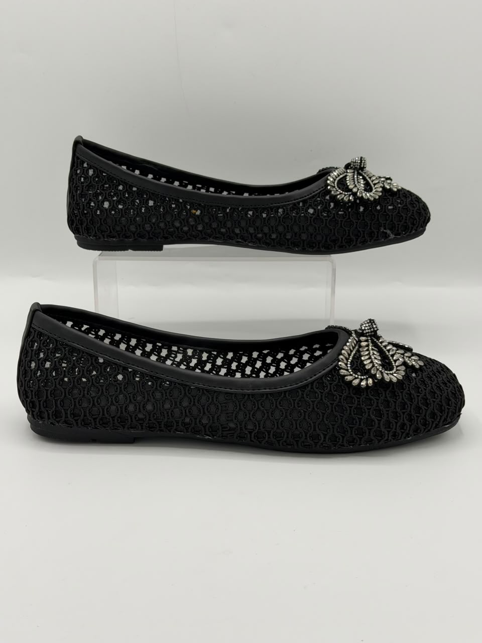 A pair of black knit ballet flats with silver rhinestone bow embellishments, displayed on a clear acrylic stand against a light gray background.