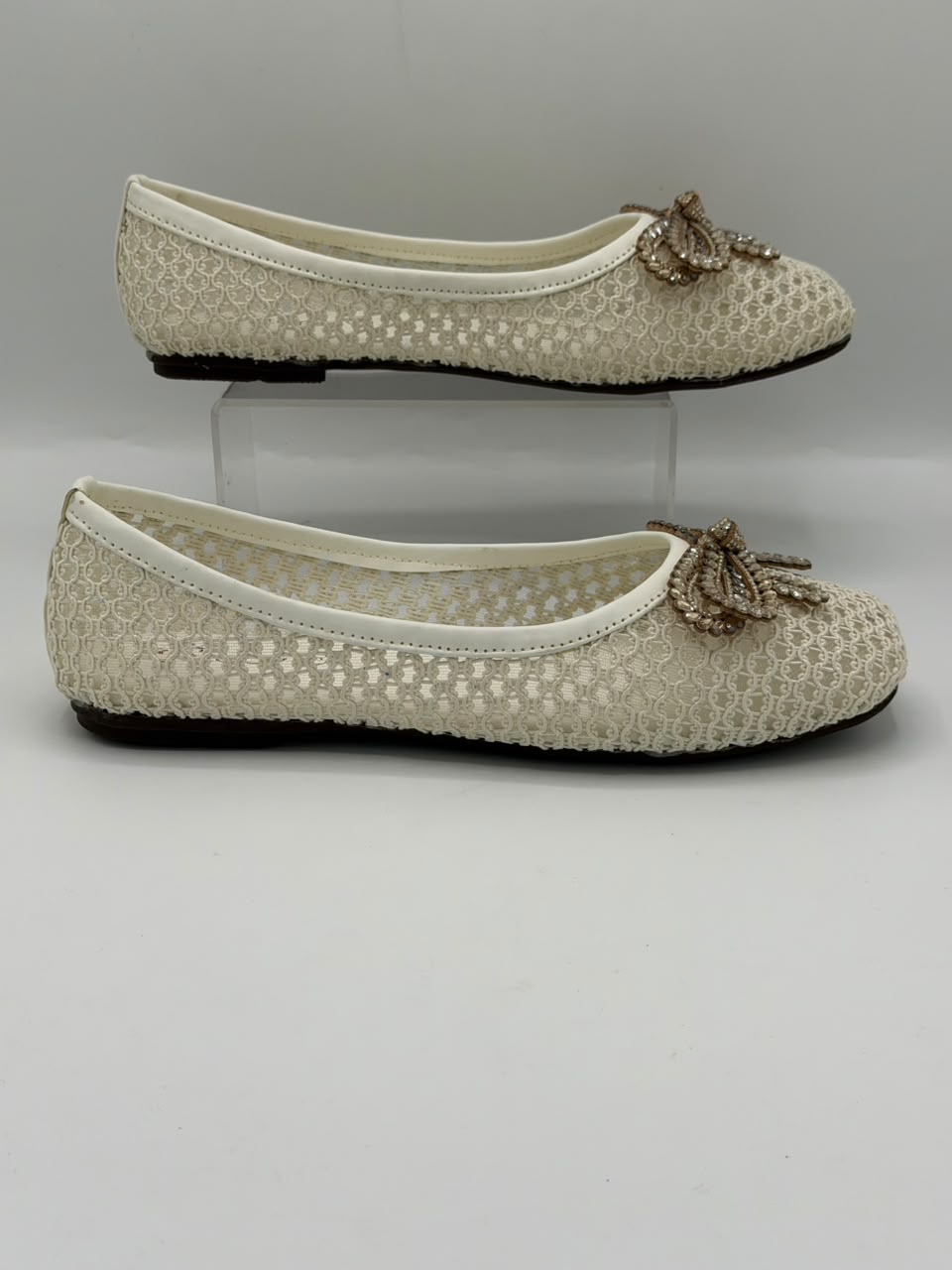 A pair of women's ballet flats in a textured cream mesh fabric, featuring a decorative metallic gold bow embellished with small rhinestones on the toe. One shoe is displayed flat on a light gray surface, while the other rests on a clear acrylic display block.