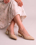A person wearing beige pointed-toe slingback pumps with chunky gold block heels, paired with a light floral skirt, seated against a pale pink studio background.
