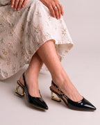 A person seated cross-legged wearing black pointed slingback heels with metallic gold block heels and a light floral skirt against a plain light gray studio background.