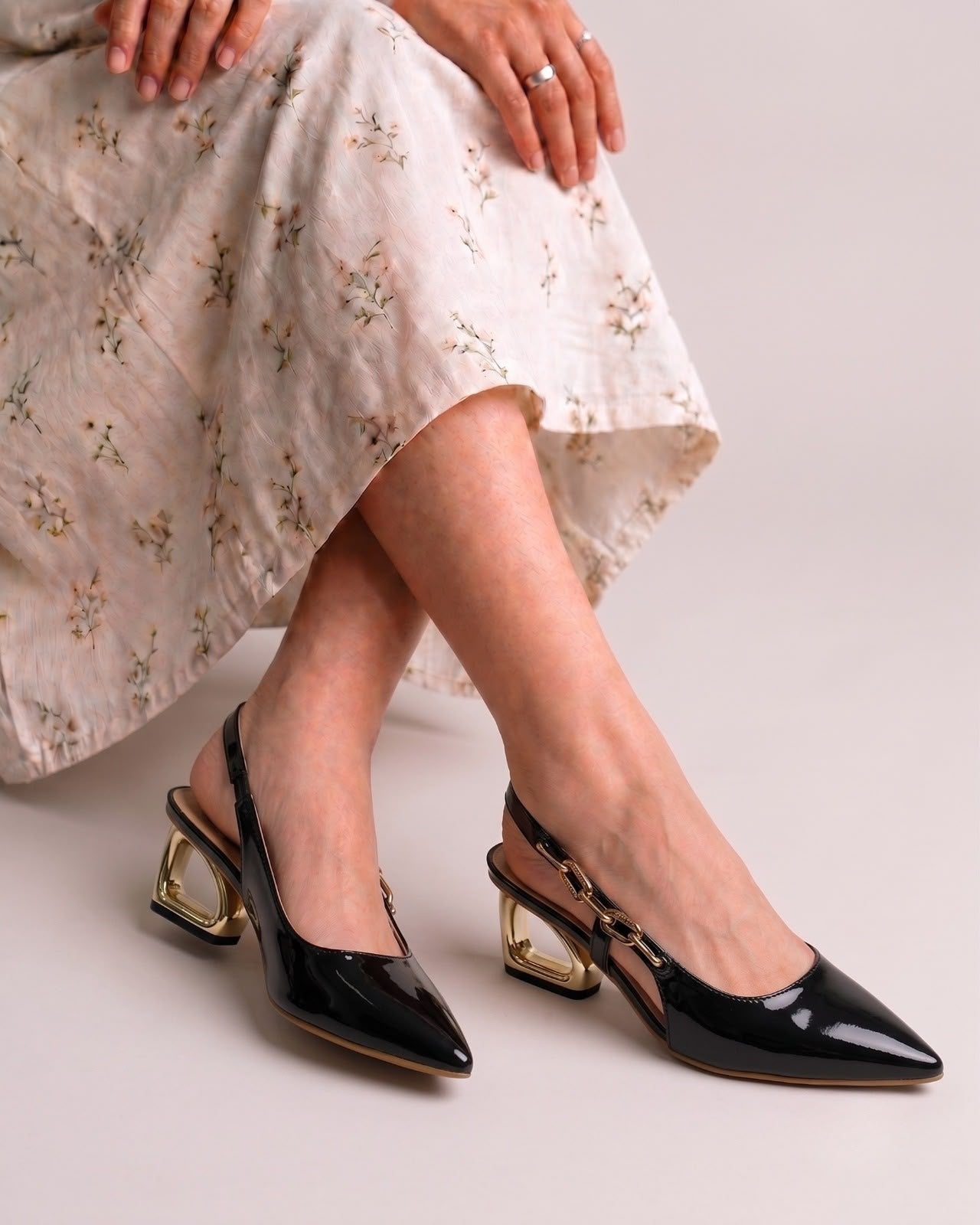 A person seated cross-legged wearing black pointed slingback heels with metallic gold block heels and a light floral skirt against a plain light gray studio background.
