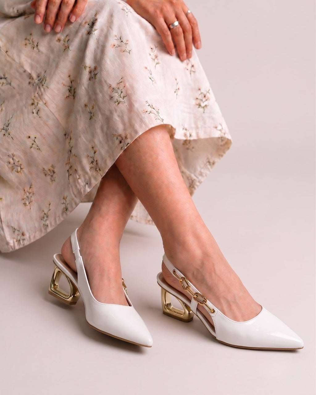 White pointed-toe slingback heels with chunky gold block heels, shown on a model wearing a light floral skirt against a neutral studio background.