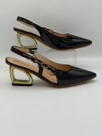 A pair of black patent leather slingback pointed-toe heels with chunky gold metal block heels, displayed on a clear acrylic riser against a light gray background.