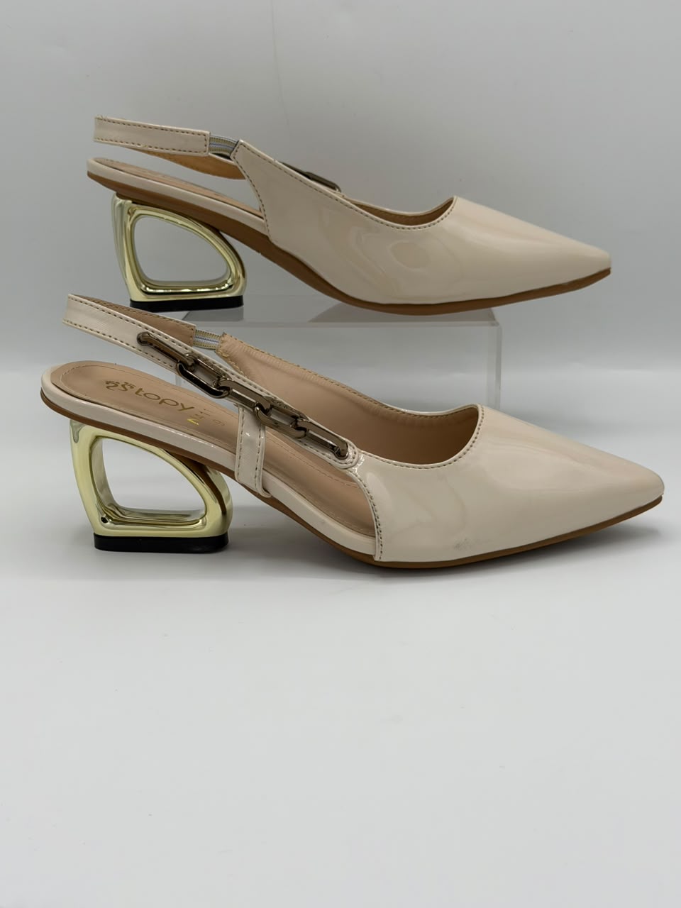 A pair of beige pointed-toe slingback heels with a unique geometric gold metal block heel, displayed on a light gray background with one shoe resting on a clear acrylic riser.