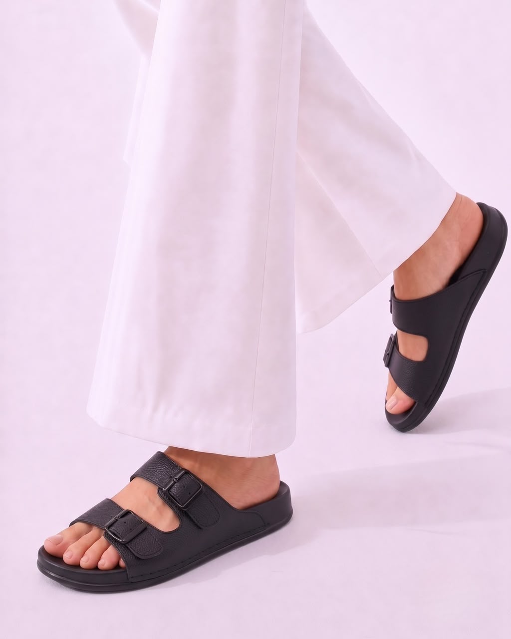 Black double-strap slide sandals worn with white wide-leg pants, shown on a model against a light lavender studio background.