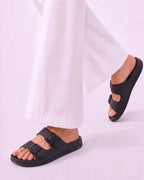 Black double-strap slide sandals worn with white wide-leg pants, shown on a model against a light lavender studio background.