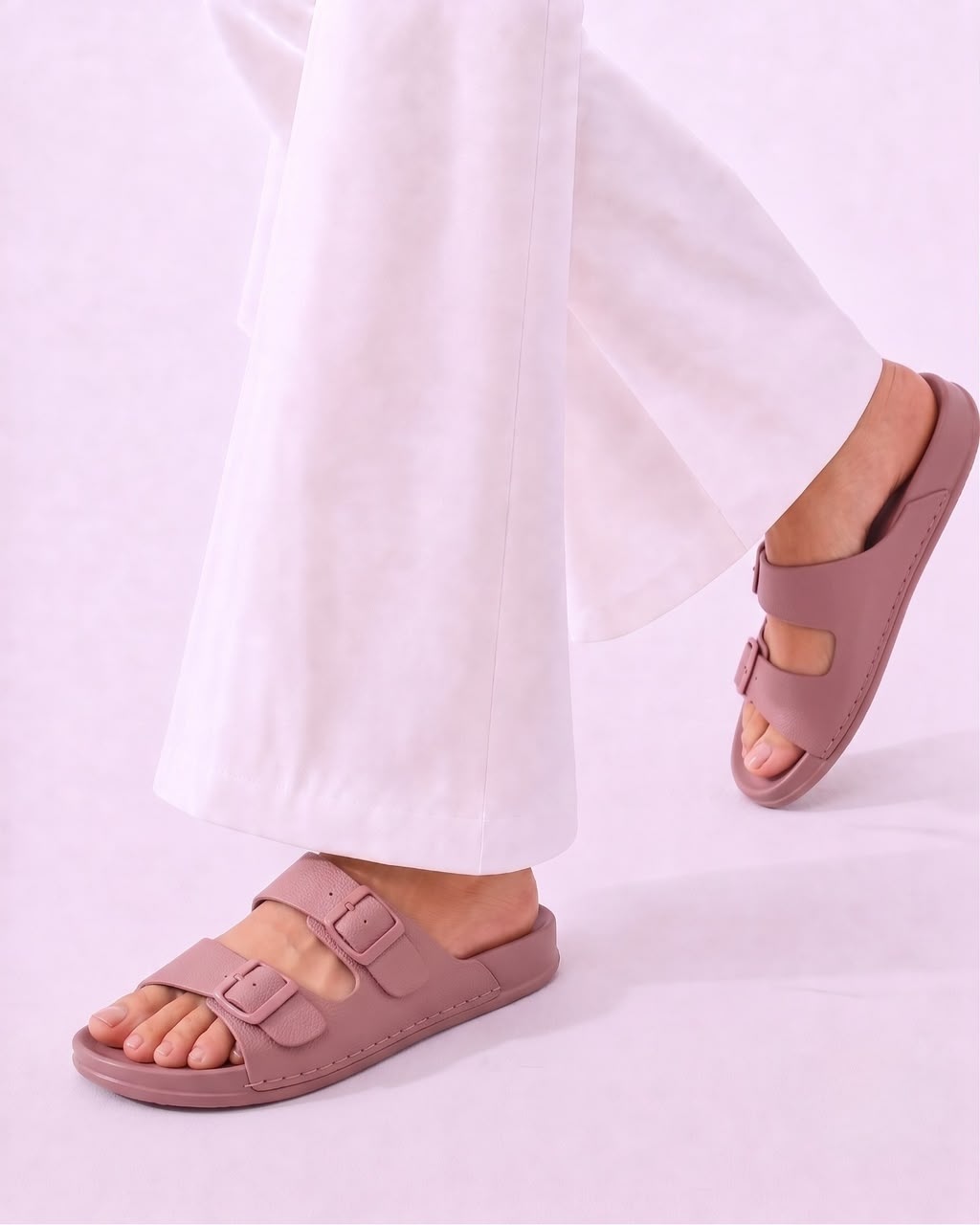 A person wearing dusty pink double-strap slide sandals with white wide-leg pants, shown from the calves down against a light lavender studio background.