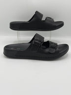 A pair of black double-strap slide sandals displayed on clear acrylic risers against a light gray background.