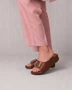 A person wearing brown leather slide sandals with large decorative square buckles, paired with dusty pink cropped pants, shown from the knees down against a light gray ribbed studio background.