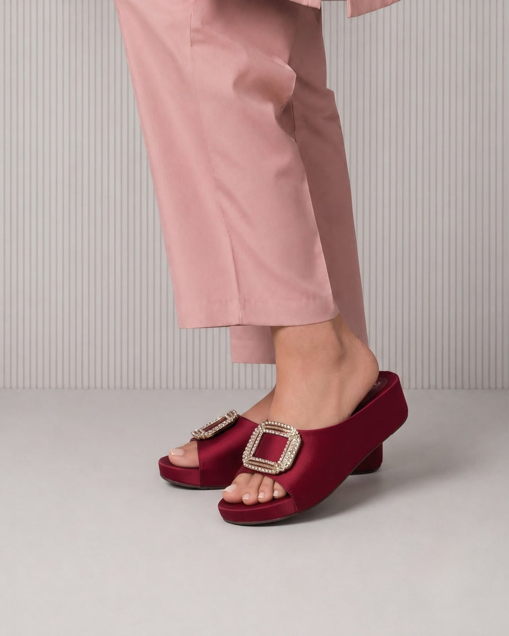 A person wearing dusty pink trousers and deep burgundy satin slide sandals with large square crystal-embellished buckles, shown from the calves down against a light gray ribbed studio background.