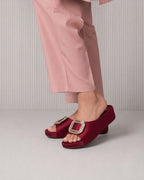A person wearing dusty pink trousers and deep burgundy satin slide sandals with large square crystal-embellished buckles, shown from the calves down against a light gray ribbed studio background.
