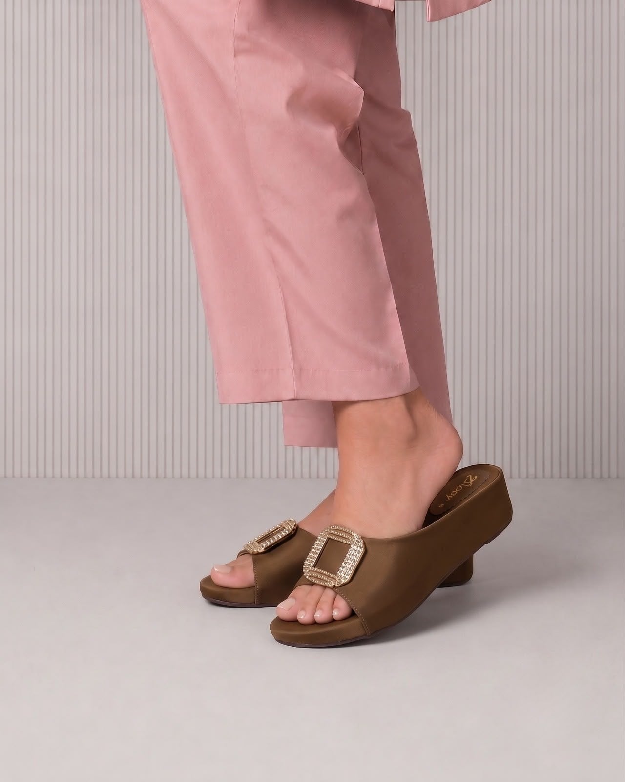 A person wearing dusty pink trousers and brown slide sandals with a prominent square buckle, shown from the waist down against a light gray ribbed studio background.