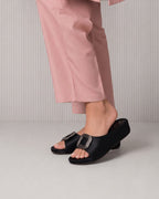 A person wearing black slide sandals with wide straps and silver rhinestone-embellished square buckles, paired with light pink trousers, shown from the knees down against a light gray ribbed studio background.