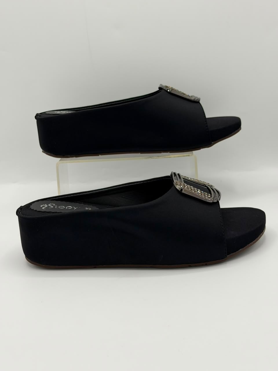 A pair of black wedge slide mules with silver-tone rhinestone-embellished square buckles, displayed on a clear acrylic riser against a white studio background.