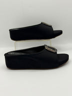 A pair of black wedge slide mules with silver-tone rhinestone-embellished square buckles, displayed on a clear acrylic riser against a white studio background.