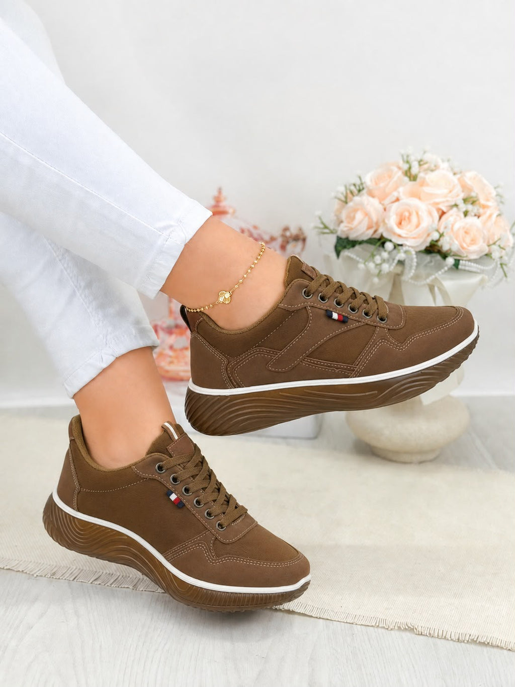 A pair of women's brown low-top sneakers with white midsoles, shown on a model's feet. The model is wearing white pants, and a decorative vase of pink roses is visible in the background.