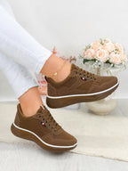 A pair of women's brown low-top sneakers with white midsoles, shown on a model's feet. The model is wearing white pants, and a decorative vase of pink roses is visible in the background.