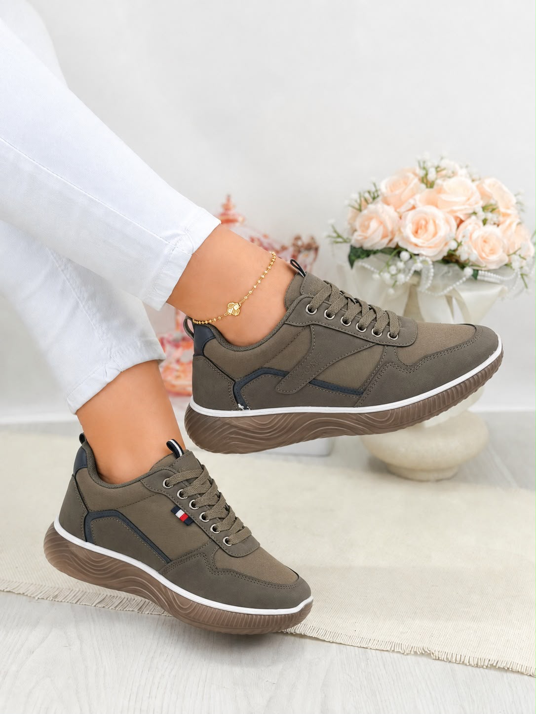 A pair of women's low-top sneakers in olive green and brown, shown being worn with white pants. The shoes feature a textured brown sole, dark brown laces, and subtle dark navy blue accents on the side panels and heel.