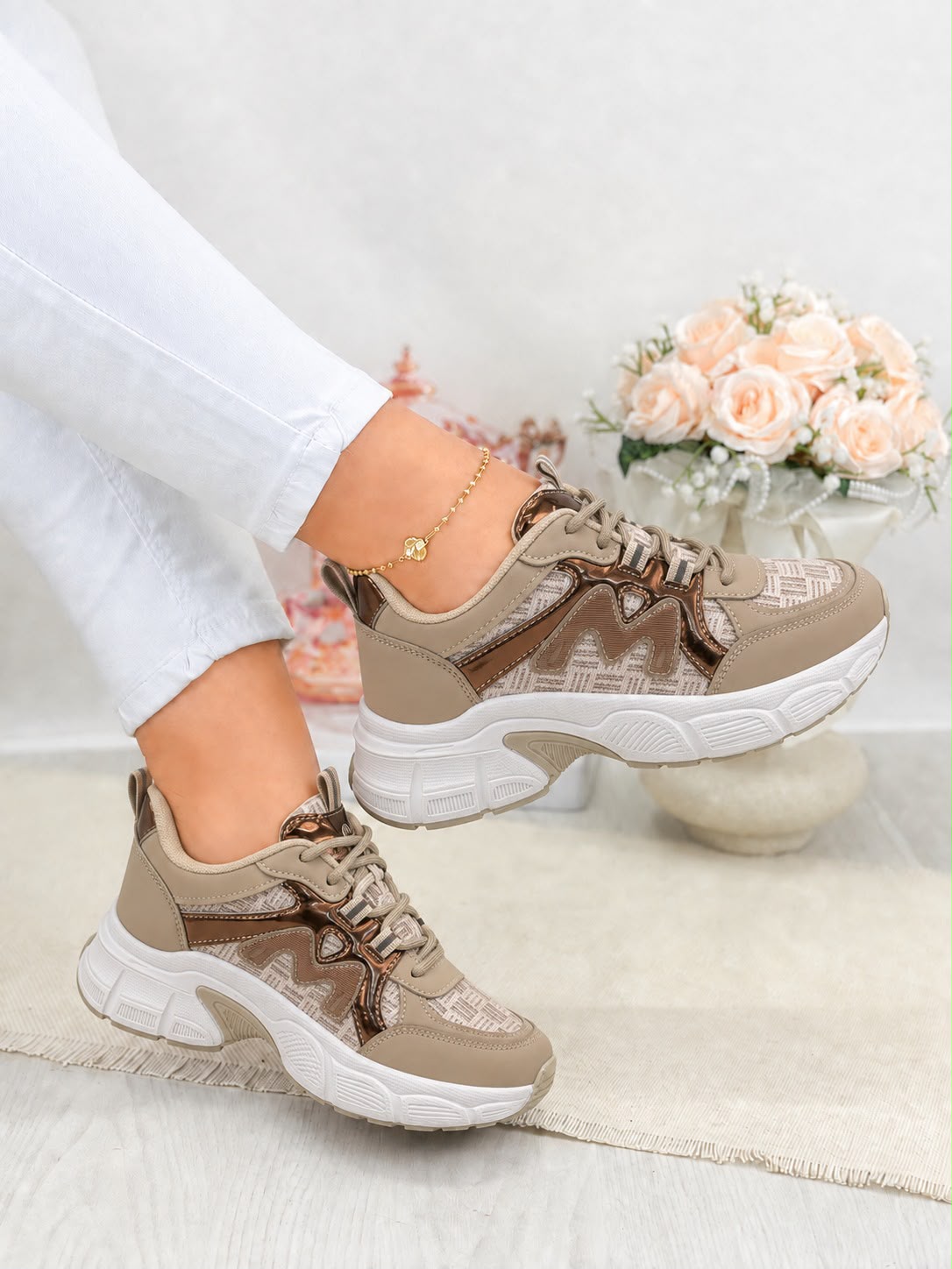 A pair of chunky low-top sneakers in beige and brown with white soles, shown on a model wearing white pants. The background features a soft-focus floral arrangement.