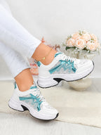 White low-top sneakers with turquoise and gray patterned side panels, shown being worn with white pants in a bright indoor setting with a vase of pale pink roses in the background.