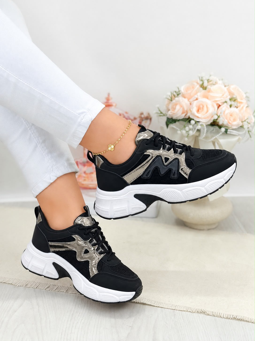 A pair of black chunky platform sneakers with metallic silver and gold geometric accents, shown on a model wearing white pants in a brightly lit indoor setting with a vase of pink roses in the background.