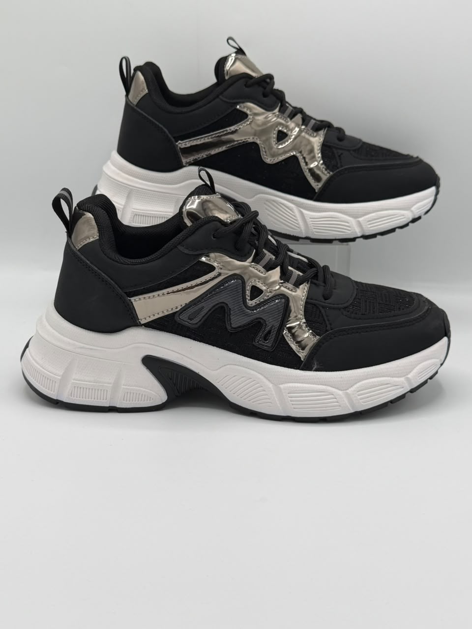 A pair of black and white chunky platform sneakers with metallic silver accents, displayed on a light gray background.