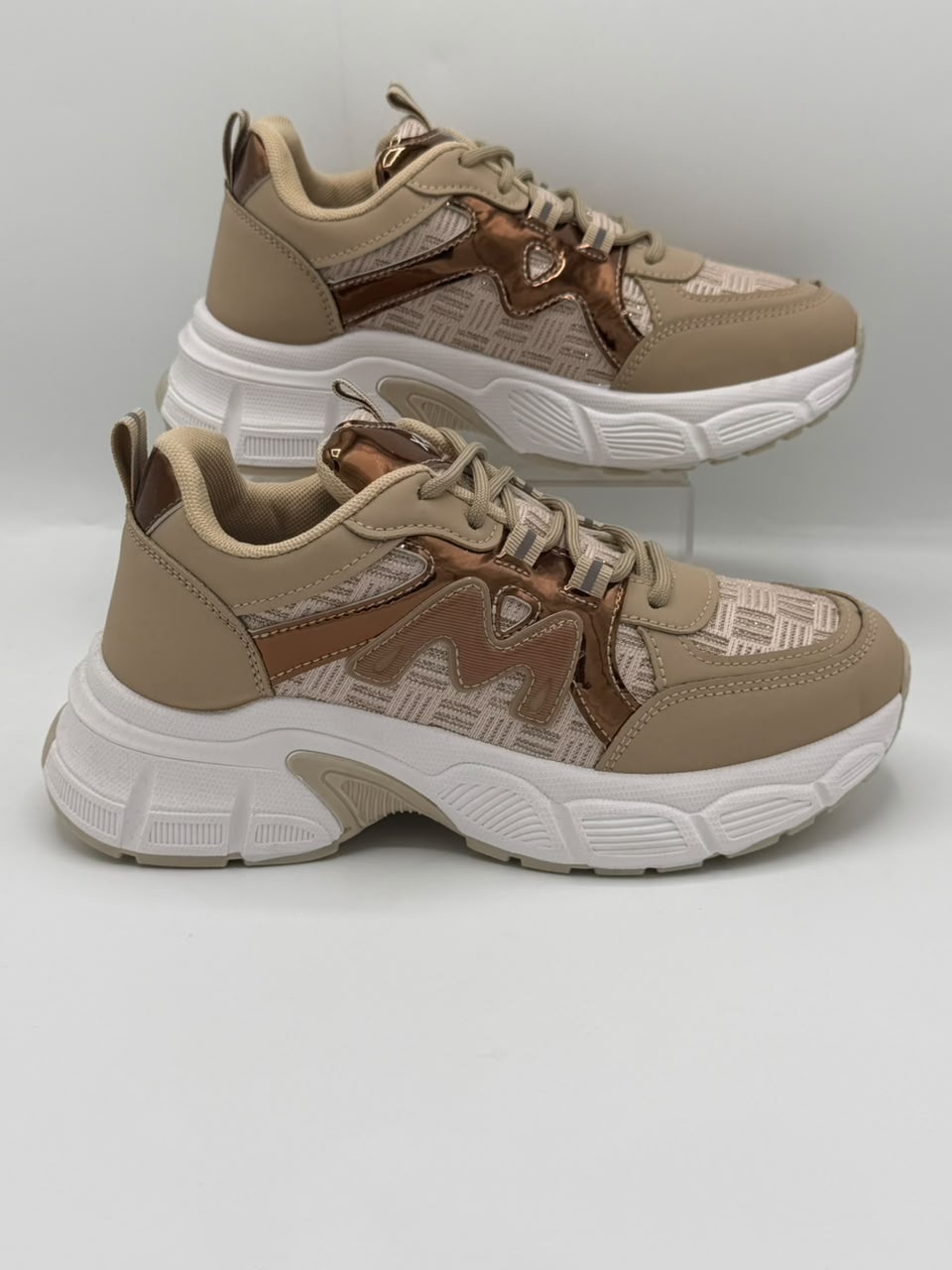 A pair of chunky low-top sneakers in beige and brown with a metallic bronze 'M' logo and a white sole, displayed on a light gray background.