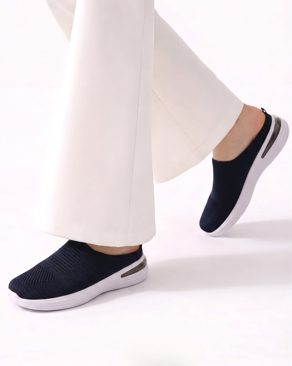 A person wearing dark navy slip-on knit sneakers with white soles and white wide-leg pants, shown from the calves down against a clean white studio background.