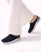 A person wearing dark navy slip-on knit sneakers with white soles and white wide-leg pants, shown from the calves down against a clean white studio background.