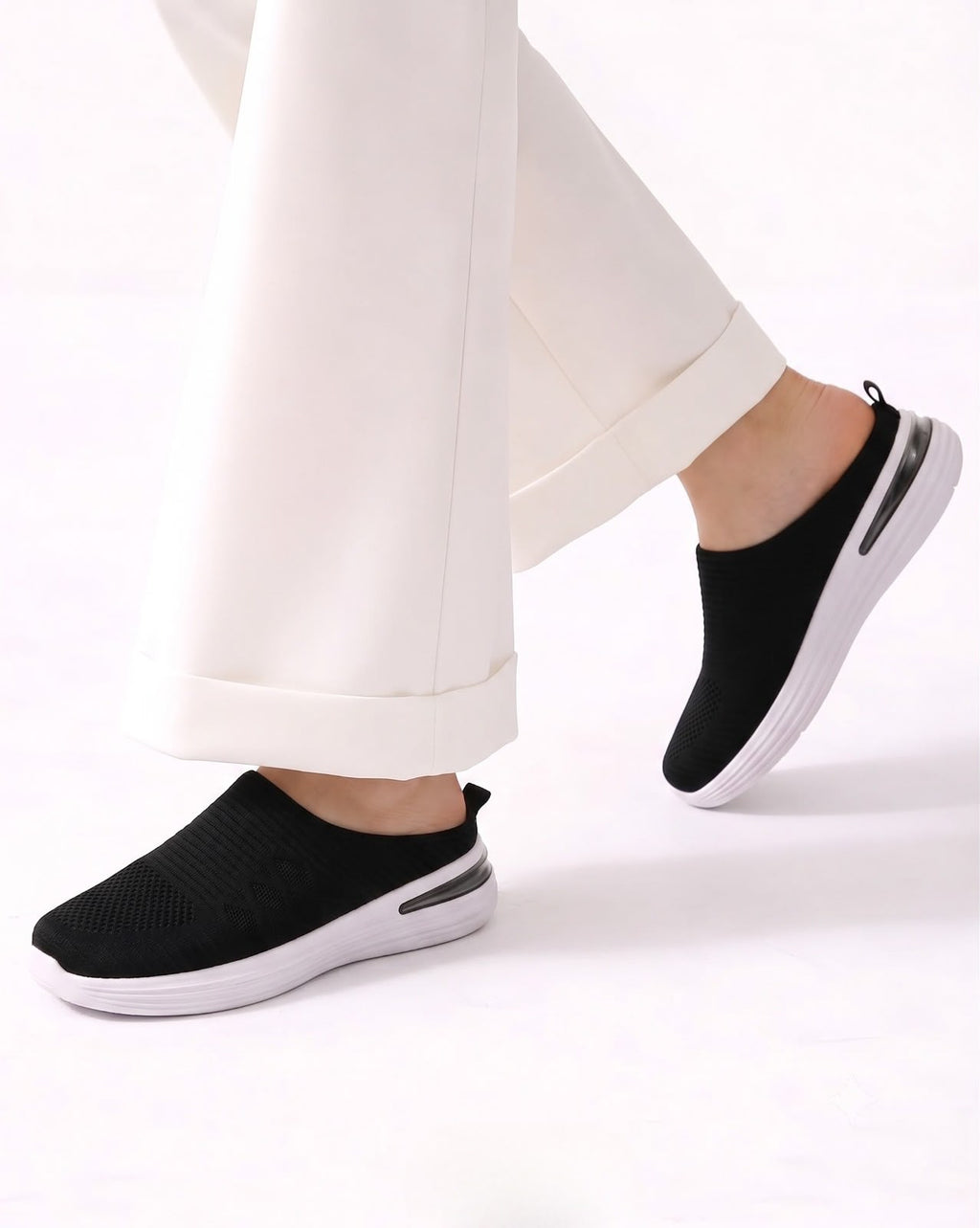 Black slip-on knit sneakers with white soles, shown on a model wearing white cuffed trousers against a clean white background.