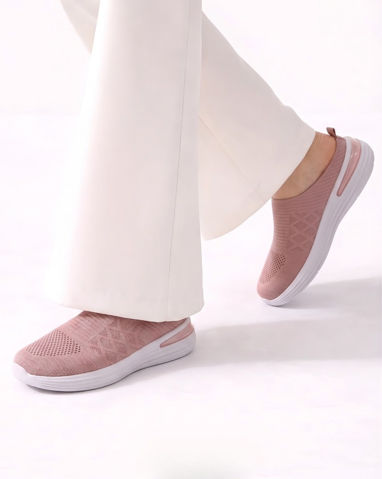 A person wearing white wide-leg pants and dusty pink slip-on knit sneakers with white soles, shown against a clean white studio background.
