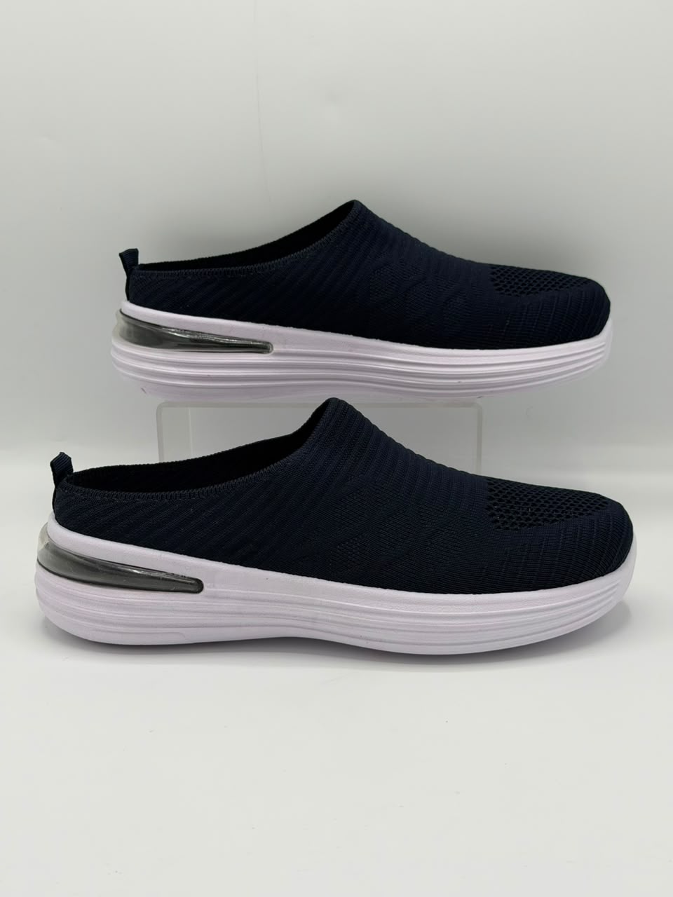 A pair of women's slip-on mule sneakers in navy blue with textured knit uppers, white soles, and a clear plastic support stand, set against a plain light gray background.