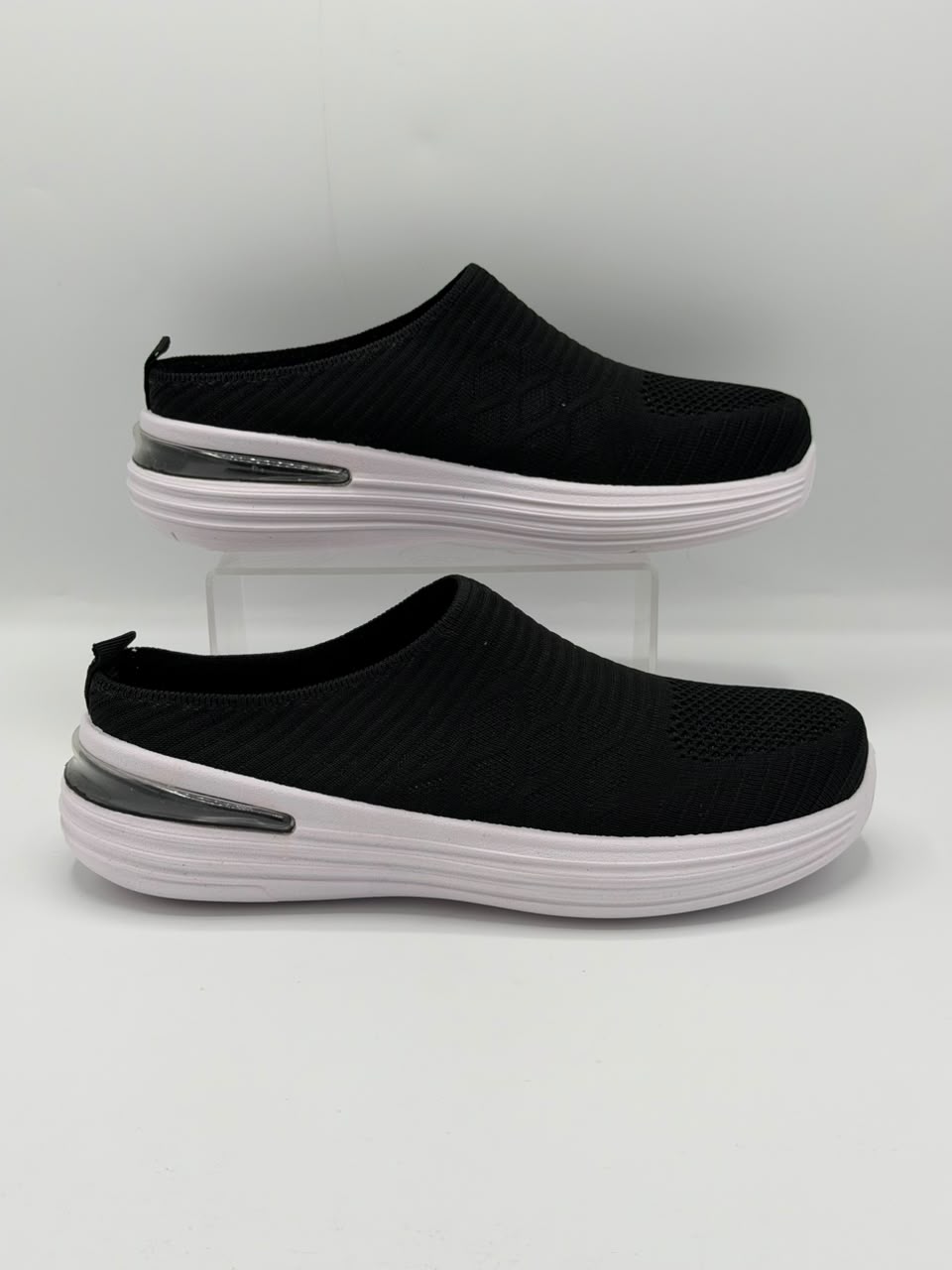 A pair of black slip-on mule sneakers with thick white platform soles and a clear heel accent, displayed on a light gray background.