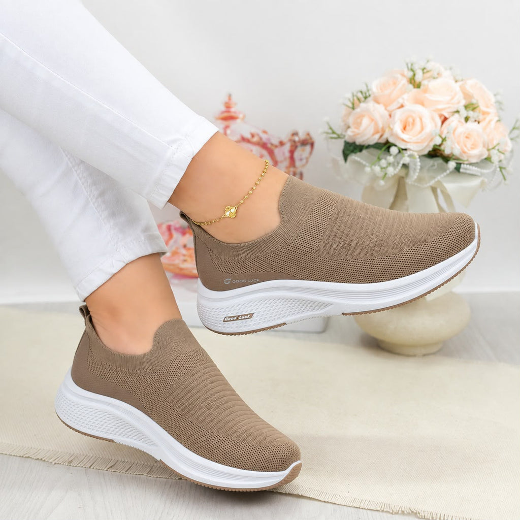 A pair of women's slip-on knit sneakers in taupe brown with thick white soles, shown being worn with white pants in a styled indoor setting with a vase of pink roses in the background.
