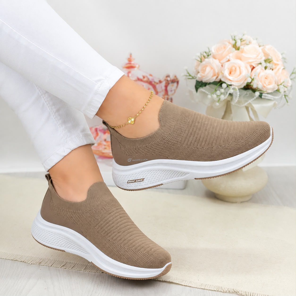 A pair of women's slip-on knit sneakers in taupe brown with thick white soles, shown being worn with white pants in a styled indoor setting with a vase of pink roses in the background.