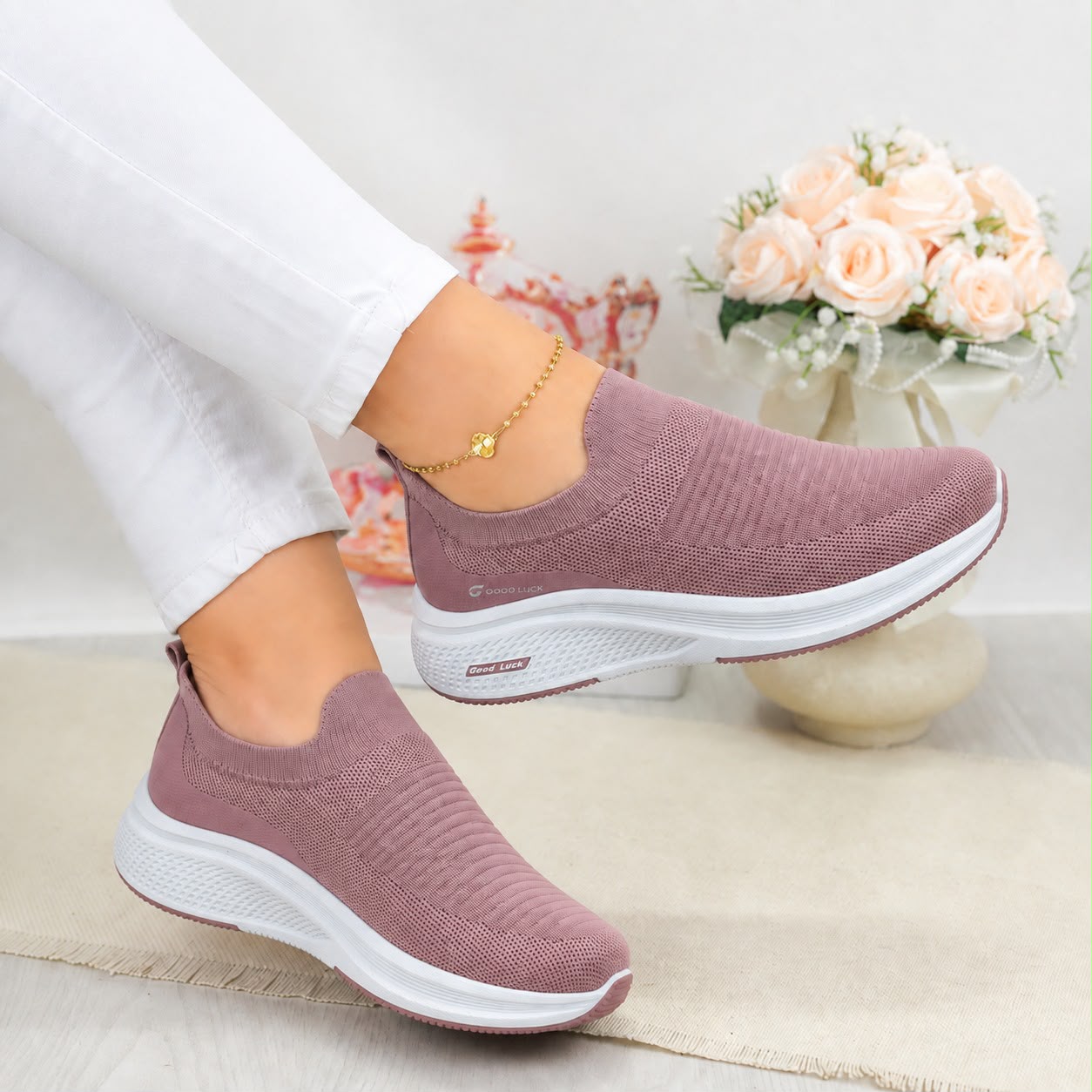 A pair of women's slip-on knit sneakers in dusty mauve pink with thick white soles, shown being worn with white pants. The background is a soft-focus indoor setting with a vase of pale pink roses.