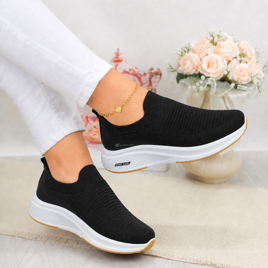 A pair of women's black slip-on knit sneakers with chunky white midsoles and a thin tan outsole, shown being worn with white pants in a styled indoor setting.