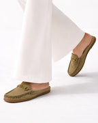 A person wearing olive green suede slip-on loafers with gold-tone horsebit details, paired with white wide-leg trousers, shown against a clean white background.