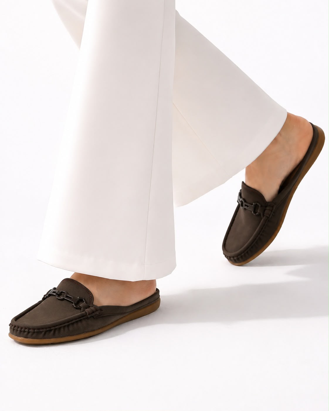 A person wearing dark brown leather slip-on loafers with chain details and white wide-leg trousers, shown from the calves down against a clean white studio background.