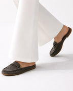A person wearing dark brown suede loafers with a metal chain detail on the vamp, paired with white wide-leg trousers, shown against a clean white studio background.