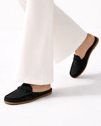 Black mule loafers with a decorative chain detail on the vamp, shown on a model wearing white wide-leg trousers against a clean white studio background.