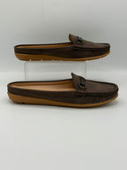 A pair of dark brown leather slip-on mule loafers with decorative tassels on the vamp, displayed on a clear acrylic riser against a light gray background.