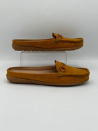 A pair of tan suede slip-on mule loafers with tassel details, one resting flat on a white surface and the other elevated on a clear acrylic block, against a white studio background.
