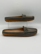 A pair of gray suede slip-on loafers with tan soles, displayed on a white background with one shoe resting on a clear acrylic riser and the other below it.