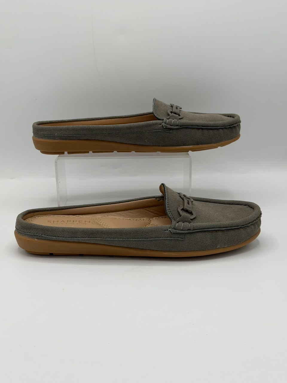 A pair of gray suede slip-on loafers with tan soles, displayed on a white background with one shoe resting on a clear acrylic riser and the other below it.